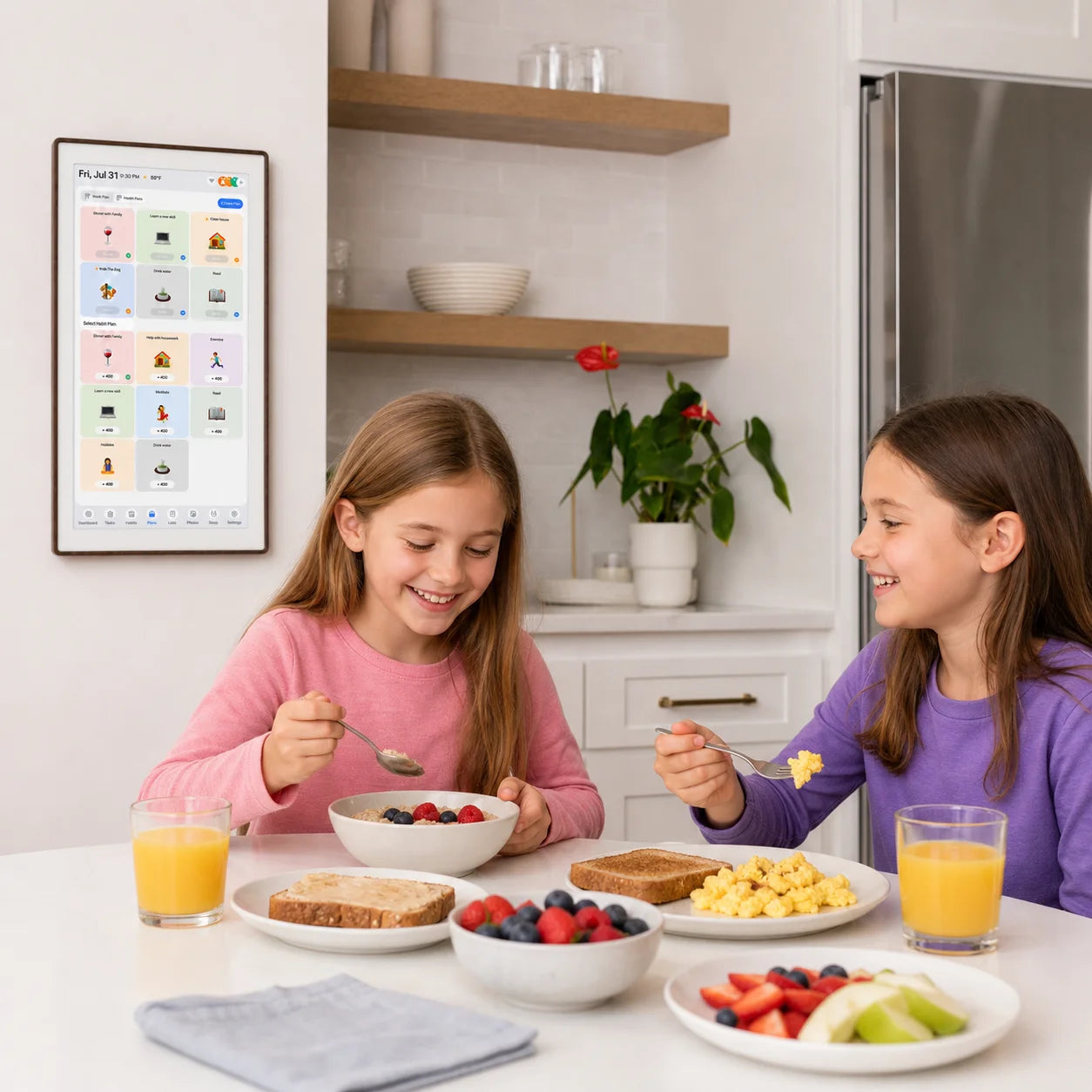 Two young girls in a kitchen looking at a wall-mounted smart screen displaying recipes or educational content, integrating digital tools into daily home life.